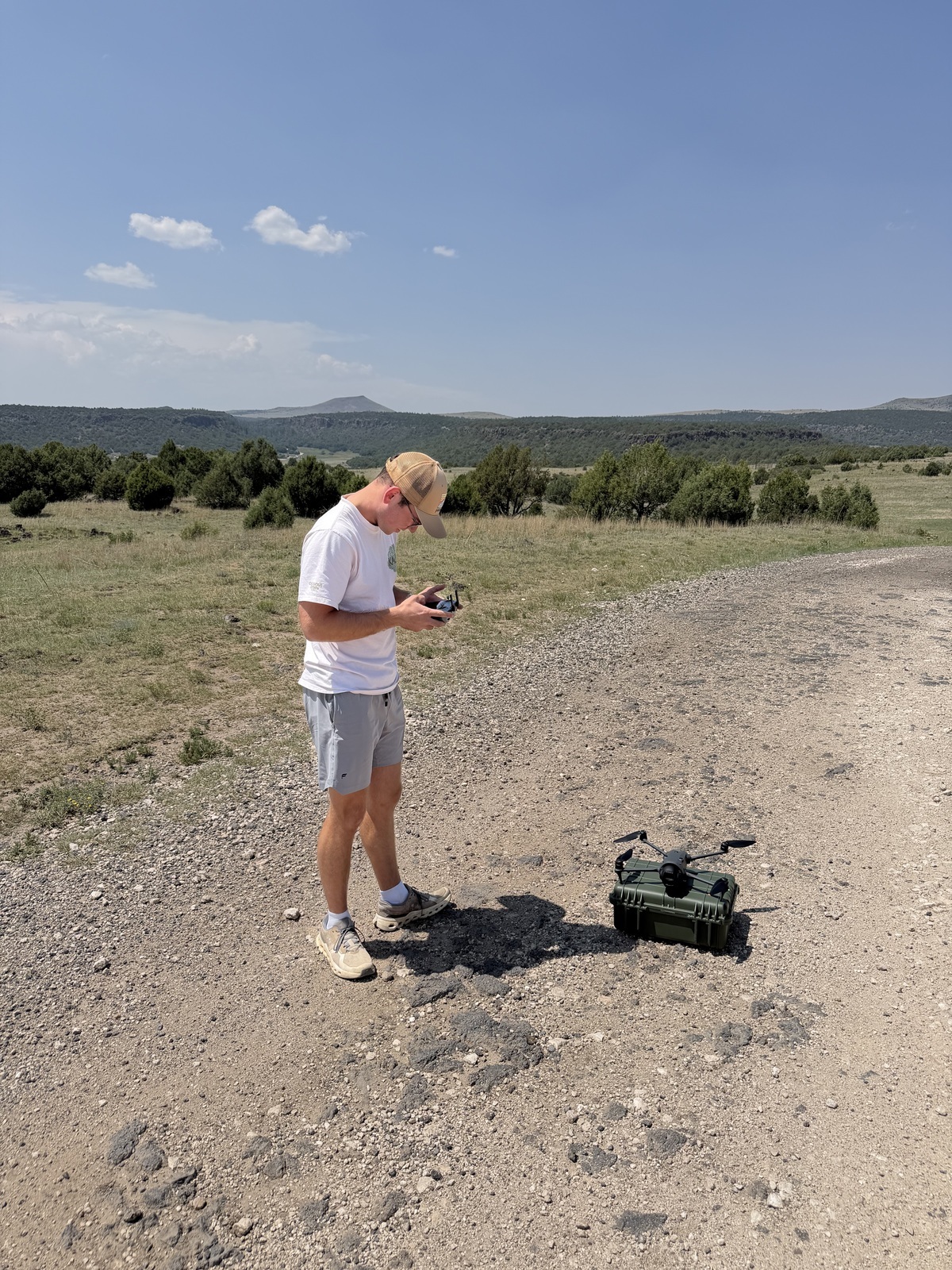 Ryan Killian standing on a gravel road in open grassland, holding a drone controller and looking down at it, with a drone resting on a hard case beside him and low mesas along the horizon under a clear sky.