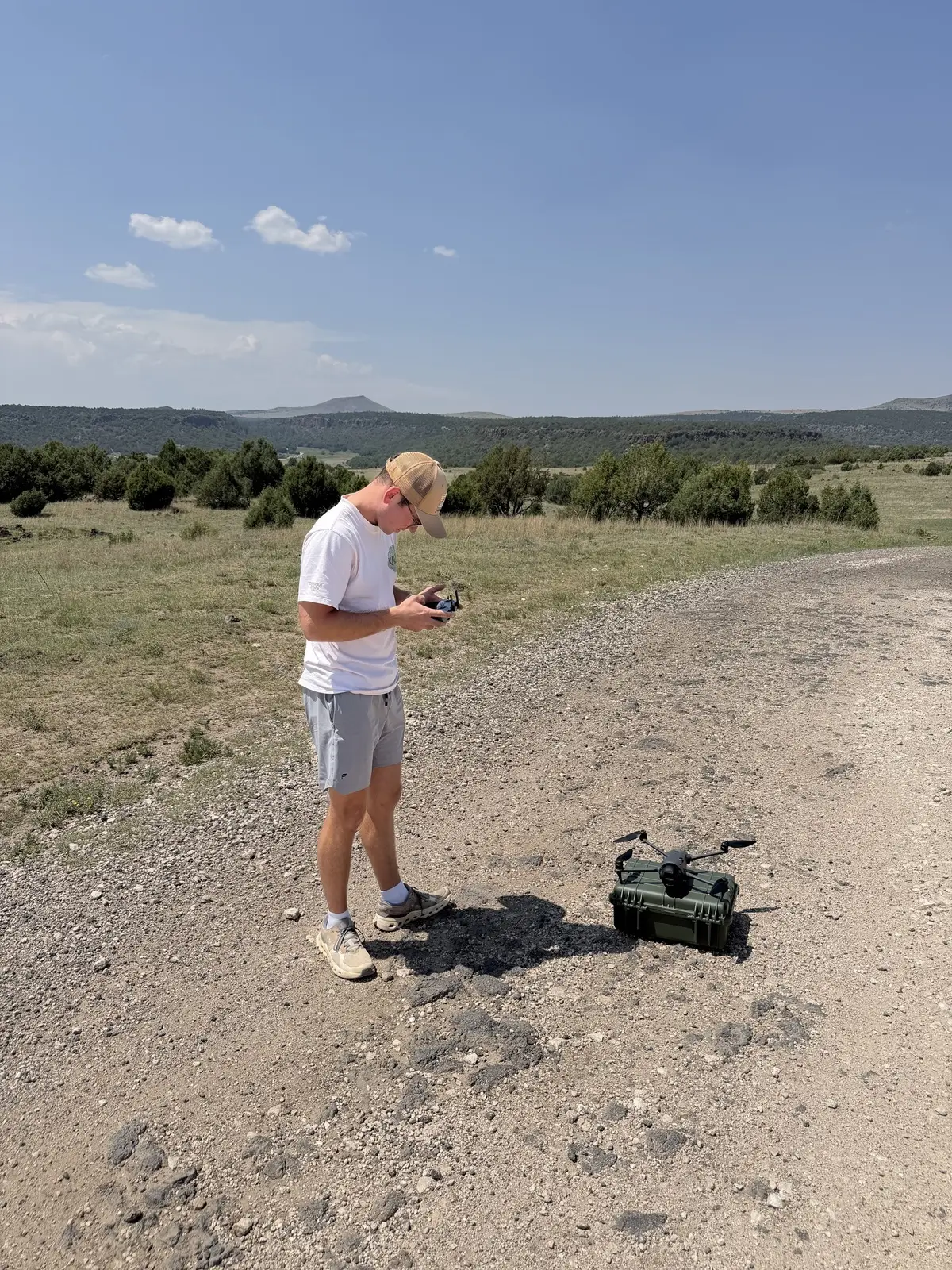 Ryan Killian standing on a gravel road in open grassland, holding a drone controller and looking down at it, with a drone resting on a hard case beside him and low mesas along the horizon under a clear sky.